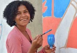 Ericka sorri em frente ao mural com pincel e tinta na mão