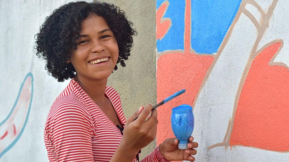 Ericka sorri em frente ao mural com pincel e tinta na mão